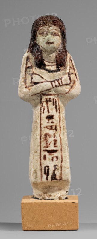 Ushabti of Mrs. As, hieroglyphs, As ca. 1295–1069 BC. Figurine in living costume, loincloth reaching to the ankles. Arms crossed on the chest, right folded over left. Hoes in both hands, basket on the back slung over both shoulders. A usekh necklace on the chest, wide bracelets on the wrists. On the head is a smooth, double wig. Round face, poorly defined nose, narrow mouth, large eyes. The wig, eyes, eyebrows, mouth, necklace, bracelets, hoes, and bag are painted in brown with a burgundy tint. The inscription is painted and runs in one vertical line on the front of the headband: Shd Osiris, Lady of the House of As. c. 1295–1069 BC; New Kingdom, 19th–20th Dynasty, Egypt. Type: shabti. Height 13.2 cm; width 4.5 cm., A small sculpted figure stands upright on a rectangular base, depicting a person with crossed arms and a stylized wig. The figure shows a mix of off-white, beige, and cream tones on its body, with darker brown and black accents on the hair and along the arms and torso. The f...