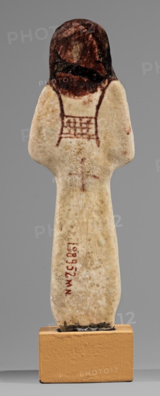 Ushabti of Mrs. As, hieroglyphs, As ca. 1295–1069 BC, usekh necklace, hoes, wigs, grave goods, Figurine in living costume, loincloth reaching to the ankles. Arms crossed on the chest, right folded over left. Hoes in both hands, basket on the back slung over both shoulders. A usekh necklace on the chest, wide bracelets on the wrists. On the head is a smooth, double wig. Round face, poorly defined nose, narrow mouth, large eyes. The wig, eyes, eyebrows, mouth, necklace, bracelets, hoes, and bag are painted brown with a burgundy tint. The inscription is painted and runs in one vertical line on the front of the headband reading: Shd Osiris, Lady of the House of As. Malgorzata Korzeniowska, c. 1295–c. 1069 BC; New Kingdom, 19th–20th Dynasty, Egypt, shabti; height 13.2 cm, width 4.5 cm, A small carved figure stands on a rectangular base, with an elongated pale body showing a mottled off-white and beige surface and a few darker speckles, topped by a rounded head that is darker brown with area...