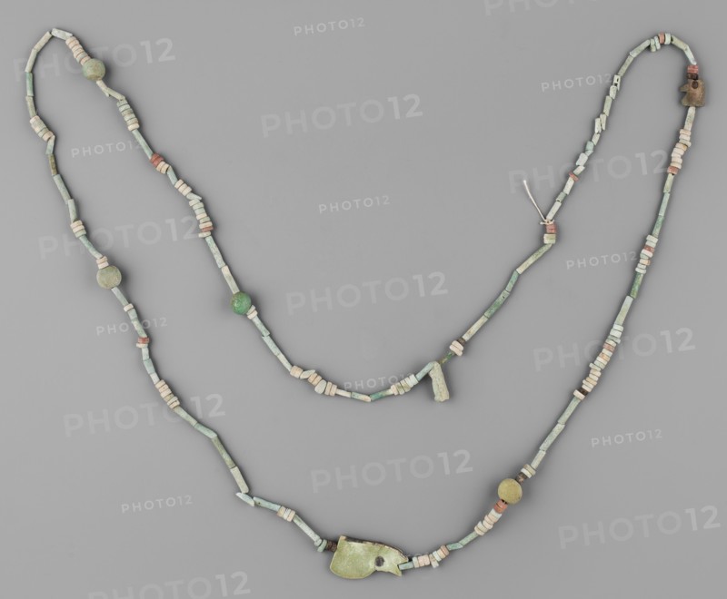 Necklace, Necklace consisting of 67 straw-shaped beads, 117 flat rings, 10 double flat rings with two holes, 4 balls, 2 udjat eyes 1 eyelet in 2 parts, 1 deity figurine without preserved feet. Predominant color: green; some beads brick-red, brown, and white. Late Period, Egypt, 664–332 BC. Amulet/jewelry; height 98 cm., A beaded necklace arranged against a neutral gray background, composed of a mix of cylindrical and disc-shaped beads and a few larger spherical beads, with a larger irregular pendant-like piece near the lower center; the beads show colors including various shades of green, pale green, off-white, beige, and touches of muted reddish-brown, with a small knot of light-colored string visible near the upper right.