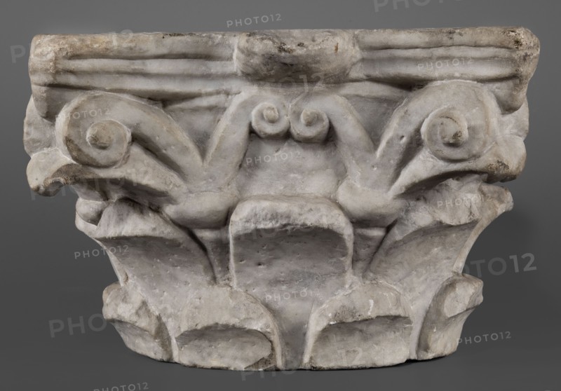 Corinthian capital, Corinthian capital composed of a calathos and thick stems that bifurcate and terminate in volutes supporting the abacus. The crown of leaves, formed by two rows of eight full leaves, occupies half the height of the capital. The leaves are rounded and angled outward. Thick caules with lacings in the form of thick rings grow from the upper row. The volute stems are in the form of flat ribbons. The outer volutes are significantly larger than the inner ones, meeting directly under the projection of the abacus rosette. The abacus is profiled, two-part with extended corners. Two corners, including the volutes, are upholstered. The edges of the leaves are chipped. Acanthus leaves ornament. 4th–5th century late antique period, Roman Empire. Architectural element; capital. Height 24 cm, width 34 cm, depth 33 cm, weight 42.5 kg., A carved stone capital with ornamental scrolls and leaf-like forms sits against a neutral background; the surface shows a weathered texture with chi...