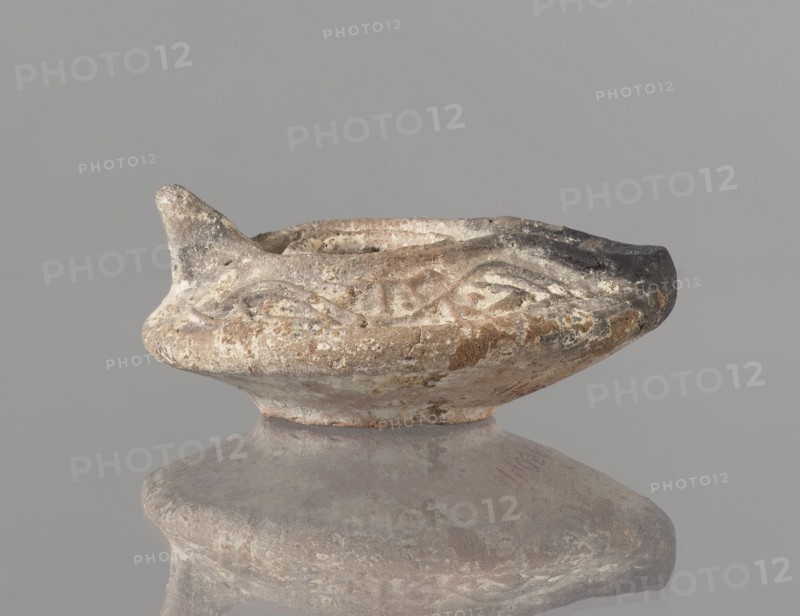 Oil lamp, Islamic art; Arabic culture; functional ceramic oil lamp. Oval lamp with short burner channel surrounded by a shaft-like disc; large oil hole occupying the entire surface of the disc. Arms decorated with plant tendrils; conical handle; ring-shaped base with a star in the center. Type A oil lamp Kubiak typology of Fustat, though similar lamps appear in Umayyad contexts late 7th–8th century. 2nd half 7th–8th century, Syro-Palestine. Height 3.3 cm; width 11.0 cm; depth 9.8 cm. Traces of use., An aged shallow bowl or lamp-like ceramic object sits on a rounded pedestal, showing worn, textured surfaces with rough edges and chips; the rim has a small pointed spout on one side and an irregular darkened area on the opposite side, with carved or incised decorative motifs around the upper body. The object displays a palette of muted earthy colors including beige, tan, brown, gray, and areas of darker charcoal, with lighter off-white encrustations and subtle mottling across its surface; ...