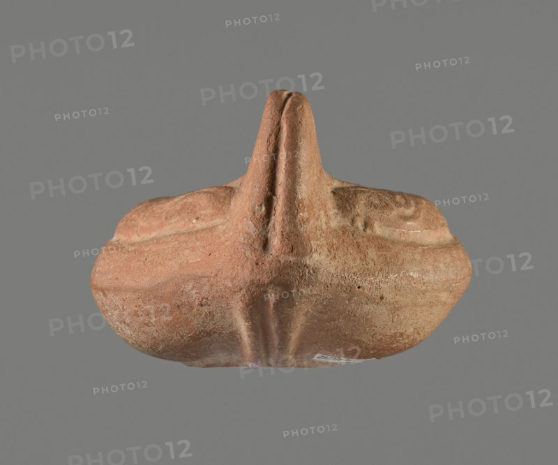 Oil lamp, Horse iconography. Lamp pressed from a mold. Light red clay with brownish-red engobe. Almond-shaped container. Ring-shaped, slightly concave base with a mold mark of two notches letter lambda or simplified alpha. A single groove runs from the base towards the vise. Solid vise. Arms decorated with a simplified winding vine s-scroll motif. Slightly concave disc with two olive holes, decorated with a depiction of a horse galloping left. Medium-length burner with an oval end and a round wick hole. No traces of use. 4th–5th century, Tunisia place of origin. Oil lamp; height 10.7 cm, width 6.9 cm., A roughly symmetrical, rounded object with a central raised ridge and a vertical protrusion, shown against a flat gray background. The surface appears textured and worn, with a coarse, sandy look. Visible colors include terracotta, muted brown, pale beige, and hints of dusty orange, with lighter worn areas and slightly darker recessed lines. The shape suggests layered contours and soft c...