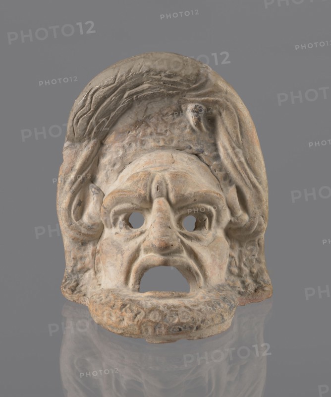 Tragic mask, terracotta, tragic masks, theater masks. Tragic mask depicting a male face. Likely a decorative element antefix?. The lower part, with the beard its underside edges are flat, is set at a near-perpendicular angle to the facial part and forms the base for the whole. A realistic representation of the appearance of a theater mask: protruding cheekbones, deep furrows around the mouth and eyes. The mouth is wide open, the lower lip almost horizontal, the upper lip arched. The large, deep-set eyeballs have circular holes cut out, positioned at slightly different heights the right one higher than the left. The nose is slightly hooked, with horizontal wrinkles. Thick eyebrows, vertical wrinkles in the center of the forehead, and a convex forehead. Above the forehead is a flat, high onkos, with schematically marked locks of hair, bounded at the top and sides by folds of cloth. Light orange clay, unevenly fired at the fracture, turning red. Traces of polychrome — red, paint on the ch...