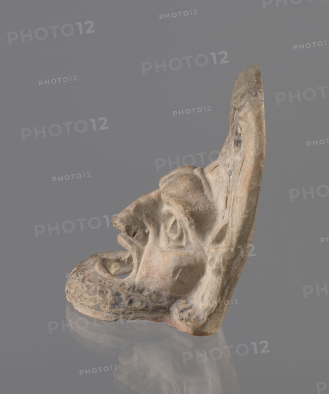 Tragic mask, terracotta, 4th–3rd century BC, Southern Italy. Tragic mask depicting a male face, likely a decorative element antefix?. The lower part with the beard underside edges flat is set at a near-perpendicular angle to the facial part and forms the base. Realistic theatrical features: protruding cheekbones, deep furrows around mouth and eyes; mouth wide open with nearly horizontal lower lip and arched upper lip; large, deep-set eyeballs with circular cut-out holes at slightly different heights right higher than left; slightly hooked nose with horizontal wrinkles; thick eyebrows, vertical forehead wrinkles, and a convex forehead. Above the forehead is a flat, high onkos with schematically marked locks of hair, bounded at top and sides by folds of cloth. Light orange clay, unevenly fired at the fracture turning red. Traces of polychrome—red, on the cheek, black, on the beard and hair. The upper right part above the forehead has been reconstructed., A sculpted fragment of a face in ...