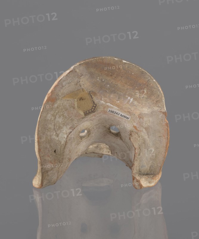 Tragic mask, terracotta, 4th/3rd century BC, Southern Italy. Tragic mask depicting a male face, likely a decorative element antefix?. Lower part with beard underside edges flat set at near-perpendicular angle to facial part forming the base. Realistic representation of a theater mask: protruding cheekbones, deep furrows around mouth and eyes; mouth wide open with lower lip almost horizontal and upper lip arched. Large, deep-set eyeballs with circular holes cut out, positioned at slightly different heights right higher than left. Nose slightly hooked with horizontal wrinkles; thick eyebrows; vertical wrinkles in center of forehead; convex forehead. Above the forehead a flat, high onkos with schematically marked locks of hair, bounded at top and sides by folds of cloth. Light orange clay, unevenly fired at fracture turning red. Traces of polychrome—red paint on cheek, black on beard and hair. Upper right part above the forehead has been reconstructed. mask, A weathered curved object with...