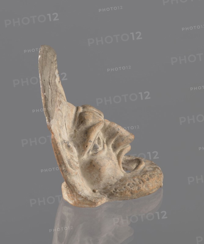 Tragic mask, terracotta, 4th/3rd century BC, Southern Italy. Tragic mask depicting a male face, likely a decorative element antefix?. The lower part with the beard underside edges flat is set at a near-perpendicular angle to the facial part and forms the base. Realistic theatrical features: protruding cheekbones; deep furrows around mouth and eyes; wide open mouth with almost horizontal lower lip and arched upper lip; large deep-set eyeballs with circular cut-out holes positioned at slightly different heights right higher than left; slightly hooked nose with horizontal wrinkles; thick eyebrows; vertical central forehead wrinkles; convex forehead. Above the forehead is a flat, high onkos with schematically marked locks of hair, bounded at top and sides by folds of cloth. Light orange clay, unevenly fired at the fracture turning red. Traces of polychrome—red, on cheek, black, on beard and hair. The upper right part above the forehead has been reconstructed., A weathered sculpted fragment...