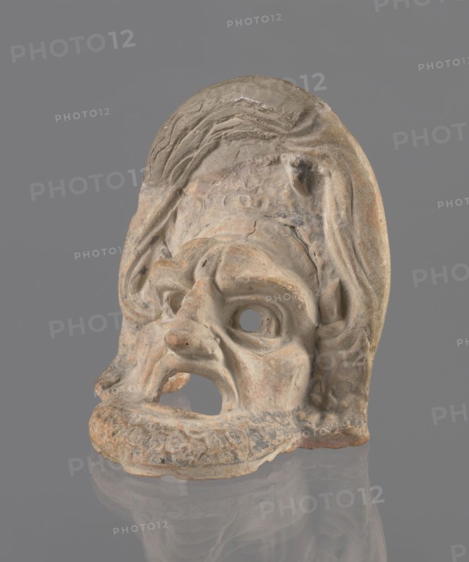 Tragic mask, terracotta, tragic/theater mask. Tragic mask depicting a male face, likely a decorative element antefix?. The lower part with the beard underside edges flat is set at a near-perpendicular angle to the facial part and forms the base. Realistic theatre-mask features: protruding cheekbones, deep furrows around mouth and eyes; mouth wide open, lower lip almost horizontal, upper lip arched. Large, deep-set eyeballs have circular cut-out holes, positioned at slightly different heights right higher than left. Slightly hooked nose with horizontal wrinkles; thick eyebrows; vertical wrinkles at center of forehead; convex forehead. Above the forehead is a flat, high onkos with schematically marked locks of hair, bounded at top and sides by folds of cloth. Light orange clay, unevenly fired at the fracture turning red. Traces of polychrome—red, on the cheek, black, on the beard and hair. Upper right part above the forehead has been reconstructed. 4th–3rd century BC, Southern Italy; mas...