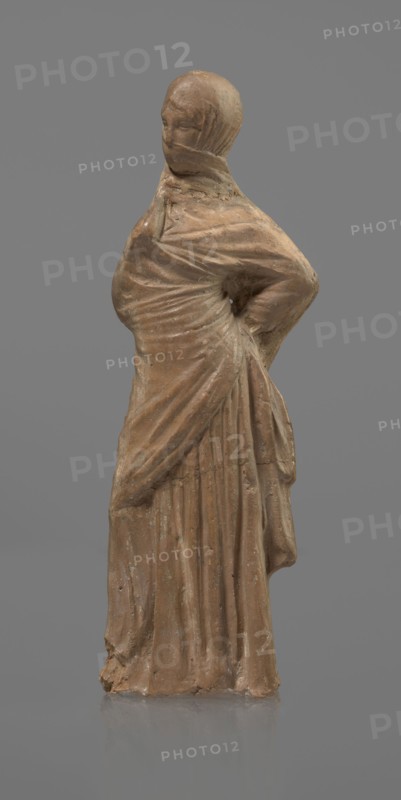 Terracotta figurine of a veiled woman, Dancers, molded female figurine, Tanagra figurine, terracotta, fine-grained red clay. Standing woman in contrapposto. Dressed in a long chiton and himation covering the head, lower face, and upper torso. Both arms bent: right hand placed in front on the breasts, left behind on the back. Probably a figure of a masked dancer. Condition: base stamped, head glued. 3rd century BC, Tanagra Greece. Sculpture. Height 15.9 cm, width 4.8 cm., A single standing figure sculpted from a brown material against a gray background, wearing a draped garment that wraps across the torso and falls in long vertical folds, with one arm bent at the waist creating a gathered area of fabric at the hip and the other arm less defined, the head covered by a close-fitting hood that obscures facial details, surface showing slight irregularities and worn areas, and visible colors including shades of brown on the figure and solid gray in the background.
