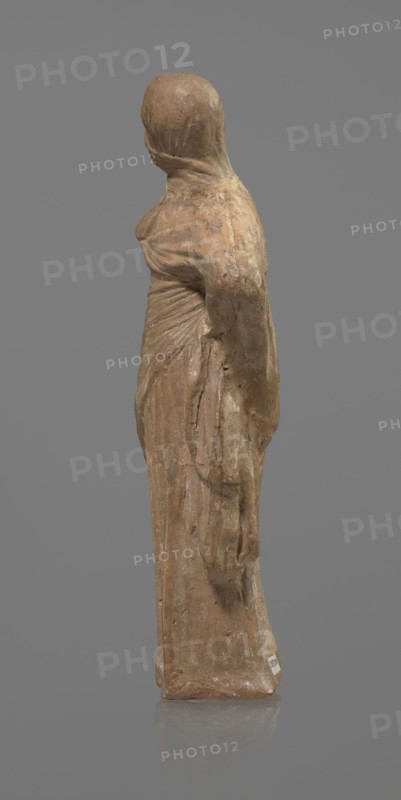 Terracotta figurine of a veiled woman, Dancers, molded female figurine in the Tanagra style, terracotta, fine-grained red clay. Figure of a standing woman in contrapposto. She wears a long chiton and a himation covering the head, lower face, and upper torso. Both arms are bent — right hand placed in front on the breasts, left behind on the back. Probably a masked dancer. Condition: base stamped, head glued. 3rd century BC, Tanagra Greece. Sculpture. Height 15.9 cm, width 4.8 cm., A worn statue of a standing figure draped in long flowing garments, shown in profile and facing left, with a smooth rounded head and a wrapped cloth over the head and shoulders; the surface shows textured weathering and small chips, and the base is flat and reflective. The visible colors are shades of brown, tan, beige, gray, and a muted clay tone, set against a solid mid-gray background.