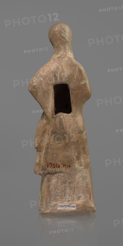 Terracotta figurine of a veiled woman, dancer, molded female figurine in fine-grained red clay Tanagra figurine. Figure of a standing woman in contrapposto. She wears a long chiton and himation covering the head, lower face, and upper torso. Both arms are bent — right hand placed in front on the breasts, left behind on the back. Probably the figure of a masked dancer. Condition: base stamped, head glued. Date: 3rd century BC. Place of origin: Tanagra Greece. Medium: terracotta sculpture. Dimensions: height 15.9 cm, width 4.8 cm., A standing figurine made of a rough textured material in shades of tan, beige, and light brown, shown from the back with a rounded head and simplified body forms; the figure has a rectangular hollow in the upper back and subtle carved folds suggesting clothing, with areas of lighter and darker tan tones and small patches of pale off-white on the surface, set against a plain neutral gray background.