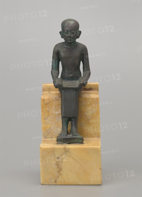 Figurine of Imhotep; seated male figure holding an unrolled papyrus on his lap, palms holding the papyrus ends. Imhotep ca. 2667–2648 BC; mythology. Materials: papyri? associated, Egyptian deities, male figurine. Date: 664–332 BC, Late Period, Egypt. Type: figurine. Dimensions: height 12 cm, width 3.2 cm., A small dark greenish bronze figure of a standing person with a smooth rounded head and simplified facial features, shown frontally with arms bent and hands holding a shallow rectangular object, wearing a vertically ribbed skirt that reaches the ankles and standing on a small rectangular base, all set on a polished yellow and cream marbled stone pedestal; the background is a uniform light gray.