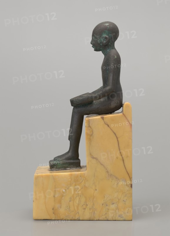 Figurine of Imhotep, Imhotep ca. 2667–2648 BC; mythology. Seated male figurine holding an unrolled papyrus on his lap, palms holding the ends of the papyrus. Material: papyri. Date: 664–332 BC, Late Period, Egypt. Height 12 cm, width 3.2 cm., A small dark bronze figurine of a seated person with a smooth rounded head and simplified facial features sits upright on a stepped block of yellowish marble with veining; the figure's arms rest on the thighs and the forearms extend forward, the legs bend at the knees with the feet placed on a lower ledge of the marble base, and patches of greenish patina are visible on parts of the bronze surface.
