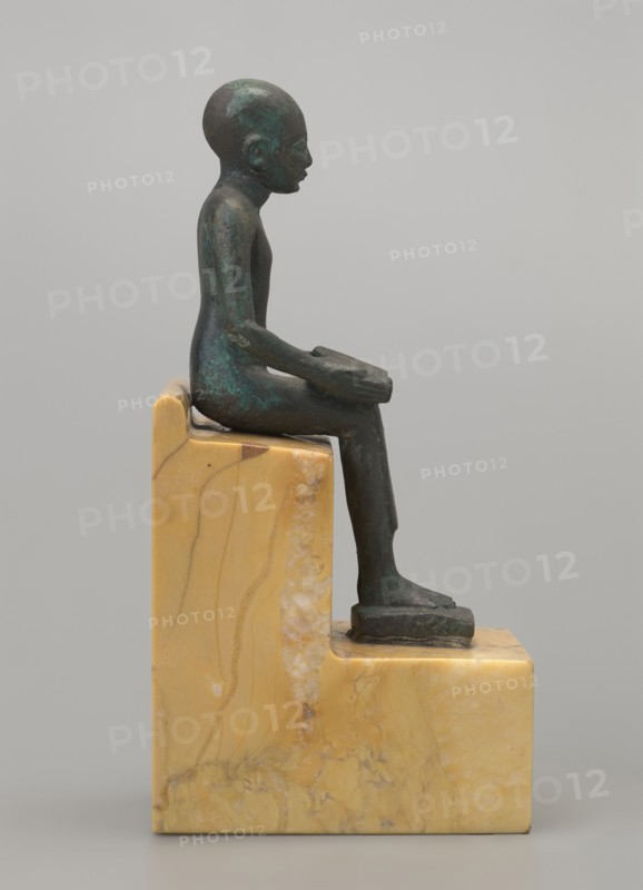 Figurine of Imhotep: seated Imhotep holding an unrolled papyrus on his lap, palms of both hands holding the ends of the papyrus. Inscribed/implied association with Imhotep ca. 2667–2648 BC; mythology. Materials: papyri depicted. Type: male figurine; style: Egyptian, Late Period. Date: 664–332 BC. Dimensions: height 12 cm, width 3.2 cm. Function: figurine., A small figurine of a seated person in a greenish dark bronze tone sits on a stepped block of yellowish beige marble with veins and patches of lighter and darker beige; the figure is shown in profile facing right with elongated limbs and a smooth rounded head, hands resting on the knees, feet placed on the lower step of the marble base, and the surface of the figure shows areas of darker and lighter greenish patina against the dark bronze.