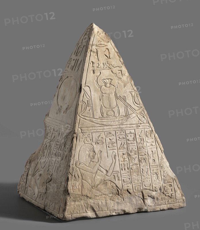 Pyramidion of a deceased named Paser and his father Turobai;, Paser ca. 1295–1186 BC and Turobai ca. 1295–1186 BC. Materials: stone architectural element; sepulchral funerary architecture; Egyptian, New Kingdom, 19th Dynasty reign of Ramesses II. Found at Deir el-Medina House no. 1312. Function: pyramidion. In the lower part Paser or his father Turobai are depicted crouching with both hands raised; surrounding free space filled with columns of hieroglyphic inscriptions. Upper register divided into four panels: I — the god Re with a human face in a sun barge; below, father or son. II — a sun barge with the Khepri scarab; below, father. III — the sign of the sun on the horizon; below, son. IV — the falcon-headed god Re with a disc on his head in a barge; below, father. Iconography includes Re, Khepri, boat, shen ring, wig, sun disc, ankh, and hieroglyphs. Dimensions: height 34.5 cm; width 25 cm; depth 23 cm; weight 16.5 kg., A carved stone pyramidion with a pointed top sits against a pla...
