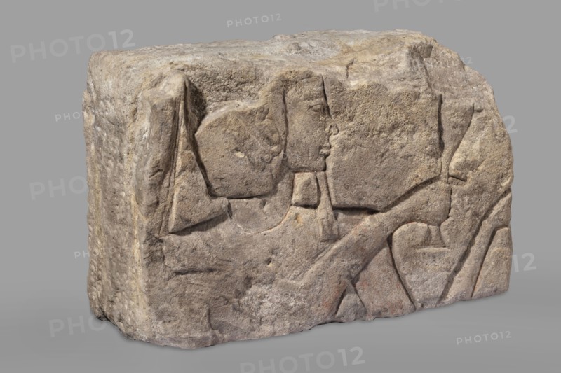 Relief depicting Ramesses II with a ritual vessel; uraeus on wig; hes vase and possible incense burner not preserved; king shown in right profile with preserved face and part of wig; wide smooth necklace around the neck; ibes wig iconographic; ritual vessels iconographic; king iconographic; religious scenes; sacred and Egyptian architecture; architectural element, bas-relief; maker, Ramesses II ruler of Egypt; ca. 1279–1213 BC, New Kingdom, 19th Dynasty reign of Ramesses II; provenance: Faras; findspot: Aksha; c. 1279–1213 BC; height 33.5 cm, width 54 cm, depth 23 cm, weight 49.5 kg., A roughly rectangular carved stone block with a weathered surface showing a carved profile of a human figure and surrounding abstract shapes; the stone appears in shades of light brown, beige, tan, gray, and hints of muted reddish tones, with textured rough and smoother worn areas, visible carved lines and shallow relief, and a neutral gray background.