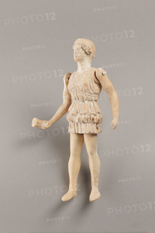 Doll with movable limbs, terracotta, made of terracotta. The doll depicts a woman in a short, pleated chiton and a bonnet; in her right hand she holds an instrument resembling castanets. The figure's oval head, directed slightly upwards, is set on a long, rather thick neck compared to the rest of the body. Lush curls protrude from under the headdress. The figurine's movable limbs are long compared to the rest of the body. 4th century BC, Attica object reported to come from Italy. Figurine, toy. Height 18.9 cm, width 8 cm, depth 3.5 cm, weight 0.13 kg., A small weathered figurine of a standing human figure with articulated shoulders and separate arms, shown against a plain neutral background. The figure wears a textured short tunic with layered folds and a belt-like section at the waist, and has short hair styled close to the head. The left arm is extended outward with a slightly bent elbow and an open hand, while the right arm hangs downward with a relaxed hand. The legs are straight w...