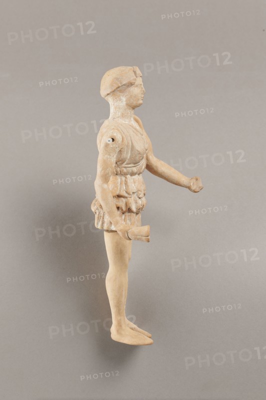 Doll with movable limbs, terracotta, dolls, Made of terracotta. The doll with movable limbs depicts a woman in a short, pleated chiton and a bonnet. In her right hand she holds an instrument resembling castanets. The figure's oval head, directed slightly upwards, is set on a long, rather thick neck compared to the rest of the body. Lush curls protrude from under the headdress. The figurine's movable limbs are long compared to the rest of the body. 4th century BC, Attica according to Weronika Krzemien the object comes from Italy. Figurine, toy. Height 18.9 cm, width 8 cm, depth 3.5 cm, weight 0.13 kg., A standing figurine viewed in profile with a light tan to beige surface showing some pale white areas, wearing a textured garment around the torso and hips with sculpted folds, a rounded head with a simple cap or hair shape, an extended arm with a hand shaped into a partial grasp, the other arm bent at the elbow holding a short cylindrical object, smooth legs and flat feet, photographed a...