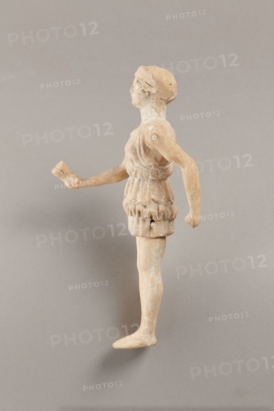 Doll with movable limbs, terracotta, dolls, Made of terracotta. The doll with movable limbs depicts a woman in a short, pleated chiton and a bonnet. In her right hand she holds an instrument resembling castanets. The figure's oval head, directed slightly upwards, is set on a long, rather thick neck compared to the rest of the body. Lush curls protrude from under the headdress. The figurine's movable limbs are long compared to the rest of the body. 4th century BC, Attica object possibly from Italy, figurine, toy, height 18.9 cm, width 8 cm, depth 3.5 cm, weight 0.13 kg., A small worn figurine of a person viewed in profile standing on a flat surface, made of a tan-beige material with patches of lighter whitish residue, wearing a short textured garment with folded and gathered details, one arm extended forward holding a small broken object and the other arm bent slightly back, a rounded head with simplified facial features and a cap-like headdress, visible joint connections at the shoulde...