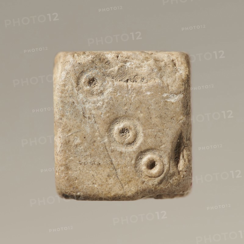Six-sided dice with pips 1–6 shown as hollow circles and dots; material unspecified; maker and place of origin, height 0.9 cm, width 1.1 cm, depth 0.9 cm., A roughly cubic object with a worn, textured surface in shades of beige, tan, and light brown, showing three recessed circular depressions on one face and slight chips and abrasions along the edges, set against a neutral light gray-beige background.