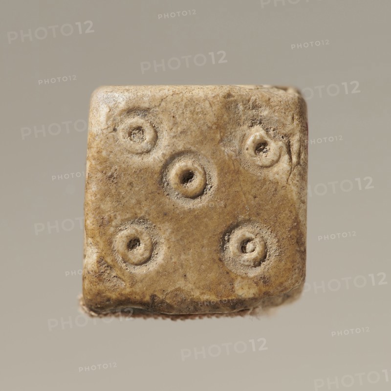 Dice, six-sided dice game, six-sided dice with points from 1–6 in the form of hollow circles and dots, unspecified material, varia, height 0.9 cm, width 1.1 cm, depth 0.9 cm, The image shows a close-up of a worn small cube with rounded edges and circular indentations on its visible face, rendered in shades of beige, tan, brown, cream, and gray, against a smooth neutral gray-beige background.