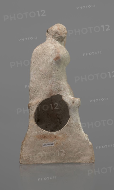 Terracotta figurine of an actor, terracotta, actors, ancient theater, male figurines. Figurine of an actor sitting on a pedestal. The head is turned to the right. The legs are crossed at the ankles. The man is wearing a loincloth and a cloak covering the left side of the body. The left arm is bent at the elbow, and the hand rests on the right cheek. The right hand holds the cloak on the left shoulder. The face is covered by a caricatured mask with a beard. Preservation condition: fragment of the cheek, left hand, and left calf is imprinted. 332–30 BC; Hellenistic period, Greece, sculpture, height 18.2 cm, A weathered sculptural object viewed from the back, showing a rounded upper form resembling a head and shoulders that transitions into a broader base with a large circular hollow in its center; the surface is rough and pitted with an aged, eroded texture. The visible colors include beige, light tan, pale pinkish tones, and patches of brown and gray, with subtle darker shading inside t...