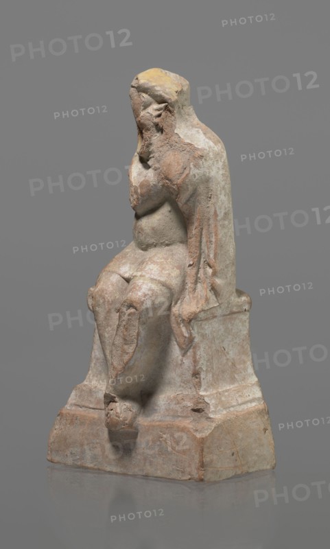 Terracotta figurine of an actor, Terracotta; Hellenistic period, 332–30 BC, Greece. Sculpture, height 18.2 cm. Figurine of an actor sitting on a pedestal. The head is turned to the right. The legs are crossed at the ankles. The man wears a loincloth and a cloak covering the left side of the body. The left arm is bent at the elbow, the hand rests on the right cheek. The right hand holds the cloak on the left shoulder. The face is covered by a caricatured mask with a beard. Preservation: fragmentary — part of the cheek, left hand, and left calf is imprinted., A weathered statue of a seated figure on a block-like base, with a draped garment over the shoulders and back, showing worn facial features and a hand raised toward the face; the surface shows rough, chipped textures and areas of erosion. The colors visible are gray, beige, tan, brown, and patches of yellow.