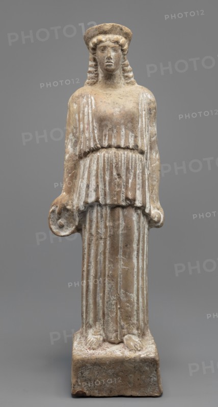 Female terracotta figurine of a standing woman positioned frontally on a quadrangular base. The figure's head, set on a massive neck, is adorned with a polos diadem. The face is oval with a large nose, parted mouth, and clearly defined eyebrow arches. Hair, pinned under the polos, falls over the shoulders and back. The body is almost entirely covered by a peplos with a long apotygm tied at the waist; the garment falls to the ground creating regular, horizontal folds. Arms fall along the torso; in her right hand she holds a patera, and in her left she clutches an object resembling an apple or pomegranate the latter supports an identification as Persephone. Traces of a chalk polychrome base are visible across the surface; remnants of light red paint are visible in the recesses of the robe and remnants of dark red polychrome on the base. Ca. 450 BC, Attica. Figurine; height 34 cm, width 10.5 cm, depth 9 cm, weight 0.75 kg., A carved standing figure of a person on a rectangular base, rende...