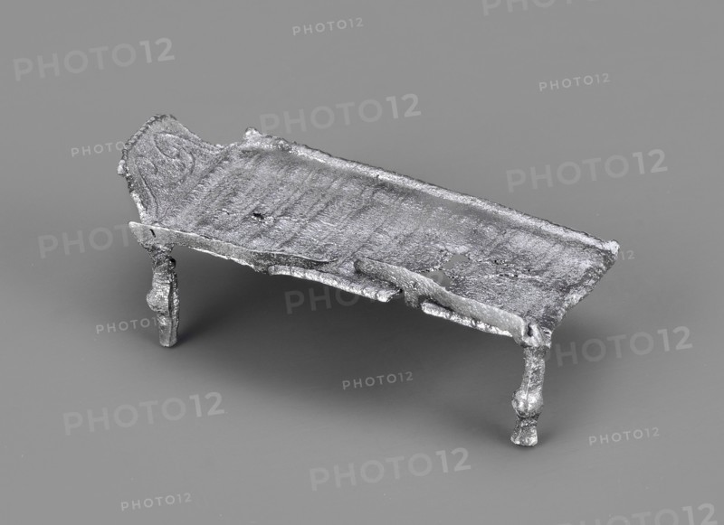 Miniature bed, votive offering or model of furniture; rests on four legs formed from square bars with spherical bulges at mid-height. The bed plate, covered with parallel slightly convex lines arranged crosswise, has long edges terminating in inwardly curled cylinders; shorter edges decorated with a row of small convex lines. A railing is attached to one long edge, mounted on four equidistant posts: two in the corners and two between them. Inside the headboard is a plastic decoration of two diagonally arranged volutes growing from the lower corners, highlighted by an arched line. 1st–2nd century Imperial Period, Roman Empire 27 BC–476, function: toy or amulet. Dimensions: height 2.1 cm, width 5.9 cm, depth 3 cm; weight 0.017 kg., A small metallic object resembling a low rectangular platform with rough, uneven surfaces and irregular edges sits on a smooth background. The platform has a textured top with visible ridges and pitted areas, and a raised section at one short end that appears ...