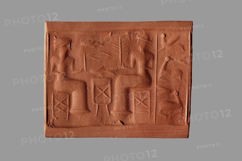 Cylinder seal with a banquet scene, unknown Assyrian workshop. The cylinder's surface is engraved with a banquet scene featuring two figures seated on either side of a sacrificial table. Above the table are two fish separated by the symbol of the goddess Ishtar — a rhombus with a line in the middle. The figures wear long smooth robes and oval-topped hats with high/wide brims, each holding a palm branch in one hand while the other arm is bent at the elbow at waist level. The vertical field behind both figures is demarcated by two vertical lines and contains three animals arranged one above the other: a winged creature on top and two antelopes lying with legs tucked under below. Iconography includes banquet scenes, rhombuses, palm branches, stools, animals antelopes, fish, and Ishtar. Eastern Gallic scene. 2nd half of the 2nd millennium BC; Middle Assyrian period, Northern Mesopotamia Assyria. Seal; height 4.2 cm, width 1.8 cm, diameter 0.3 cm opening; narrows in the middle to 0.2 cm., A...