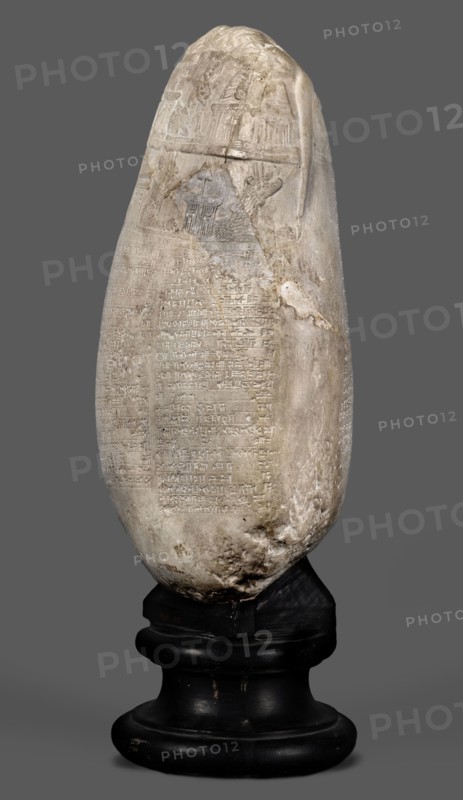 Copy of a Babylonian kudurru from the period of Marduk-naddin-ahhe 11th century BC, Ancient Near East, kudurru copy, Kassite Kingdom 1595–1158 BC, Eastern Gallic scenography, Babylonia Iraq applies to the original, 19th century, copy, weight 5.44 kg, A tall, elongated stone object with a rounded top and an irregular, weathered surface sits on a dark, rounded pedestal; the stone is mostly pale beige and off-white with areas of darker gray and brown discoloration and visible scratches and chips, and there are a few shallow carved panels and low-relief motifs near the upper portion while the lower portion shows a textured, pitted area; the pedestal is black with a slightly glossy finish and shows some worn patches and small scuffs; the background is a flat medium gray.