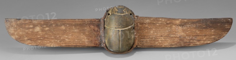 Winged scarab; protective amulet for mummies, wings, grave goods; sacred animal motif; scarab amulet made in Egypt, Late Period, 664–332 BC; materials/technique not specified; dimensions: height 5 cm, width 23.8 cm, depth 1.8 cm., A horizontal wooden object with a central carved beetle-like form attached to a pair of elongated wing-like wooden panels; the central carved form is smooth with rounded contours and shows shades of green and brown with areas of darker patina, while the wing-like panels are textured with visible grain lines and show various tones of brown and tan with lighter dusted spots; the background is a neutral gray and there is a soft shadow beneath the object.