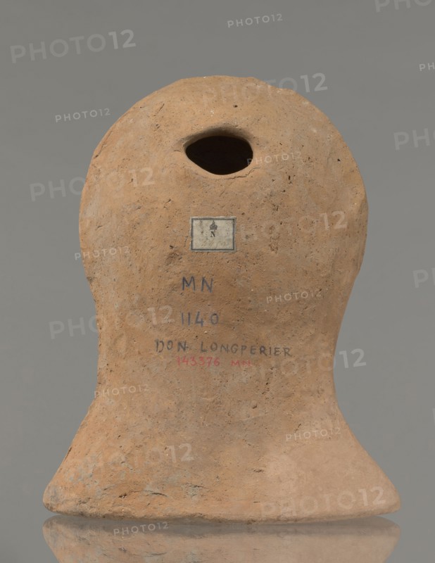 Votive female head, Female head wearing a wreath, from under which lush locks of hair curl down the sides of the face to the neck. High forehead, large eyes, deep-set in relation to the nose, slightly convex with developed eyelids and eyeballs, with pupils marked. Small straight nose, narrow slightly parted mouth with fleshy lips. Strong chin slightly protruding forward; slim cheeks giving the face a triangular outline. 3rd–2nd century BC, Etruria; votive. Height 26 cm, width 21 cm, depth 13 cm, weight 2 kg., A rounded earthen object with an irregular oval opening near the top sits against a neutral gray background; its surface is rough and pitted with a natural clay brown color and areas of slightly darker brown and pale tan; a small rectangular label is affixed below the opening and there are traces of dark blue and muted red markings on the front surface; the base flares outward into a slightly uneven foot with similar brown and tan tones and the object casts a subtle reflection on ...