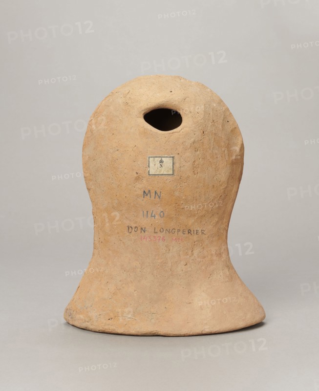Votive female head, Female head wearing a wreath, from under which lush locks of hair curl down the sides of the face to the neck. High forehead, large eyes, deep-set in relation to the nose, slightly convex with developed eyelids and eyeballs, pupils marked. Small straight nose, narrow slightly parted mouth with fleshy lips. Strong chin, slightly protruding forward. Slim cheeks giving the face a triangular outline. 3rd–2nd century BC, Etruria. Votive. Height 26 cm, width 21 cm, depth 13 cm, weight 2 kg., A rounded, bell-shaped clay object sits upright on a neutral background, showing a warm tan and light brown surface with a rough, slightly pitted texture and subtle darker speckles; the top has an irregular oval opening leading into a dark hollow, and there is a small rectangular patch of different color affixed near the upper middle; the base flares outward into a broad, uneven rim with faint variations of beige and dusty orange, and the overall form displays minor surface imperfecti...