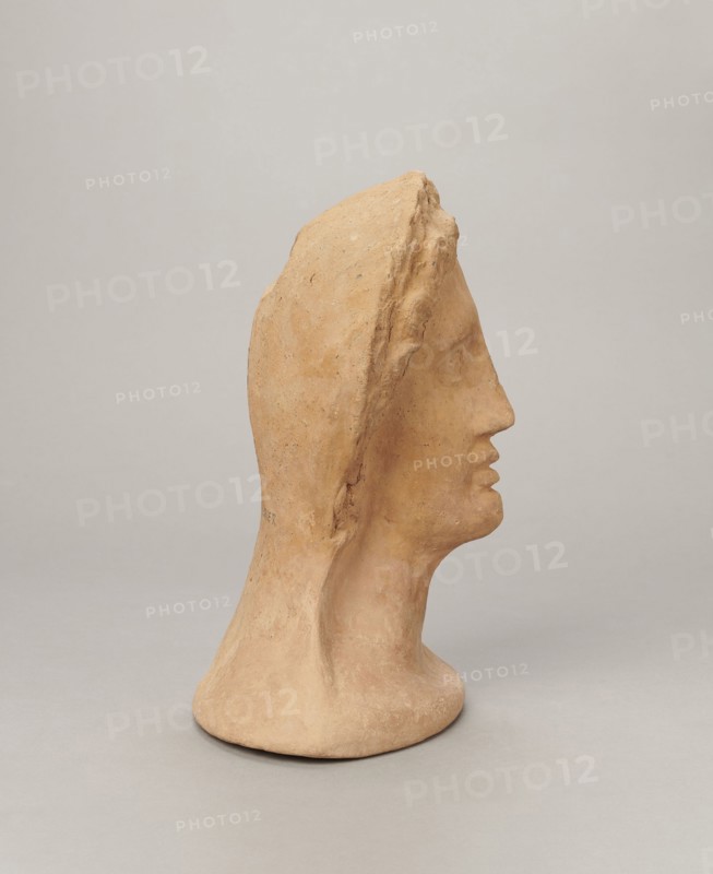Votive female head, Female head wearing a wreath, from under which lush locks of hair curl down the sides of the face to the neck. High forehead, large eyes, deep-set in relation to the nose, slightly convex with developed eyelids and eyeballs with marked pupils. Small straight nose; narrow, slightly parted mouth with fleshy lips. Strong chin, slightly protruding forward; slim cheeks giving the face a triangular outline. 3rd–2nd century BC, Etruria; votive. Height 26 cm, width 21 cm, depth 13 cm, weight 2 kg., A sculpted bust in a warm beige clay color shown in profile facing right, with a smooth elongated neck and stylized facial features including a pronounced nose and slightly parted lips, a hood-like form covering the top and back of the head with a rougher textured ridge along its edge, subtle surface variations and small speckles in the clay, and a neutral pale gray background.