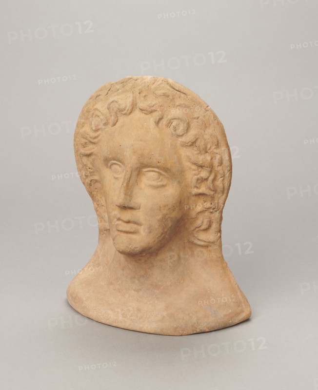 Votive female head, female head wearing a wreath, from under which lush locks of hair curl down the sides of the face to the neck. High forehead, large eyes set deep in relation to the nose, slightly convex with developed eyelids and eyeballs, pupils marked. Small straight nose; narrow, slightly parted mouth with fleshy lips. Strong chin, slightly protruding forward; slim cheeks giving the face a triangular outline. 3rd–2nd century BC, Etruria; votive. Height 26 cm, width 21 cm, depth 13 cm, weight 2 kg., A terracotta bust of a human head and neck sits against a plain light gray background, showing a stylized face with wavy hair framing the forehead and sides; the sculpture's surface is a warm earthy tan with subtle variations of darker and lighter brown, and there are hints of muted orange and pale gray in worn areas, while the background remains mostly pale gray.