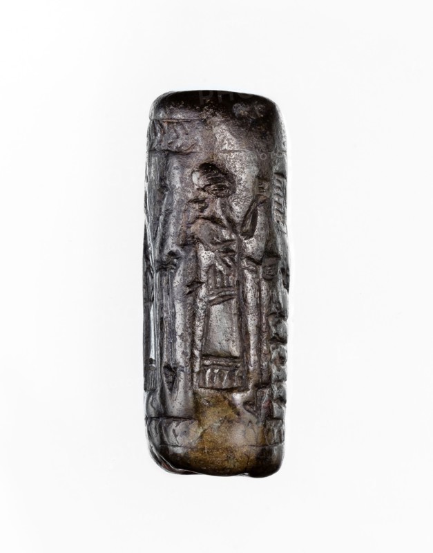 Cylinder seal, unknown Neo-Assyrian workshop. Seal in the so-called linear style with a representation of a religious-magical scene of adoration of the holy tree in the form of a stylised pomegranate bush with a winged solar disc shown above. On both sides of the bush stand human figures in profile, dressed in identical long robes with fringes at the bottom. On the left is a priest, wearing a hat with a ribbon, on the right a king and a courtier, both without headgear. In the field behind the figures are symbols: a palm leaf and an eight-pointed star. The entire image is framed at the top and bottom with a herringbone ornament. Iconography: pomegranate bushes, winged disc, stars. Linear style. Religious scenes. 9th–8th century BC; Neo-Assyrian period, Northern Mesopotamia place of manufacture; Assyria/Mesopotamia/Iraq/Turkey/Syria origins/places of finding. Cylinder seal; height 3.5 cm, width 1 cm., A vertically oriented cylindrical object with an uneven, carved surface showing a few s...