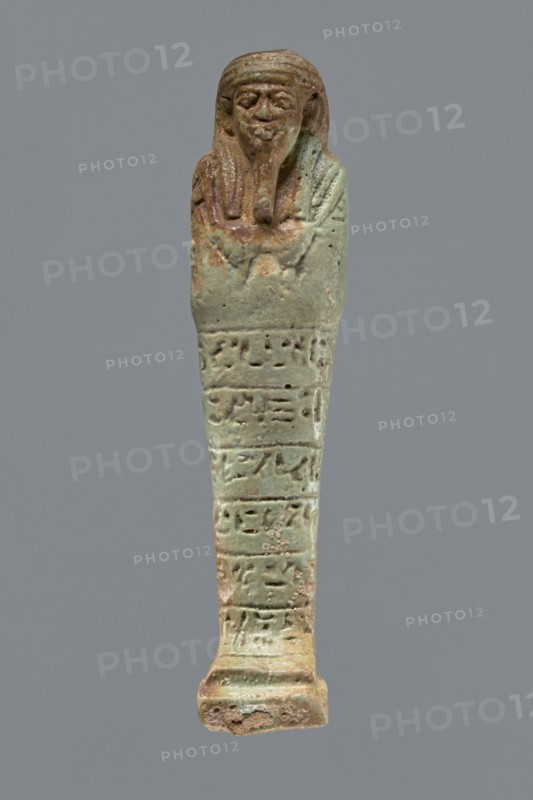 Ushabti, Mummy-shaped figurine, funerary shabti, 664–332 BC Late Period, Egypt. Standing on a base, supported by a rear pillar. Arms crossed on the chest with only the hands protruding from under the shroud, holding hoes in both hands. A basket is slung on a single strap over the left shoulder on the back. Engraved tools. Wears a three-part wig styled in narrow strands. Long, tapering beard braided and curled at the bottom. Square face with almond-shaped eyes, pronounced eyebrows, a wide nose, and prominent lips in a slight grimace. Engraved inscription arranged in six lines around the figurine; text illegible. Height 15.5 cm., A small elongated figurine with a humanoid head and a wrapped body, shown against a plain gray background, featuring a weathered surface with shades of green, turquoise, brown, and tan; the head has stylized facial features and a beard, the upper portion shows draped elements and a textured surface, and the lower portion tapers to a rectangular base with surface...