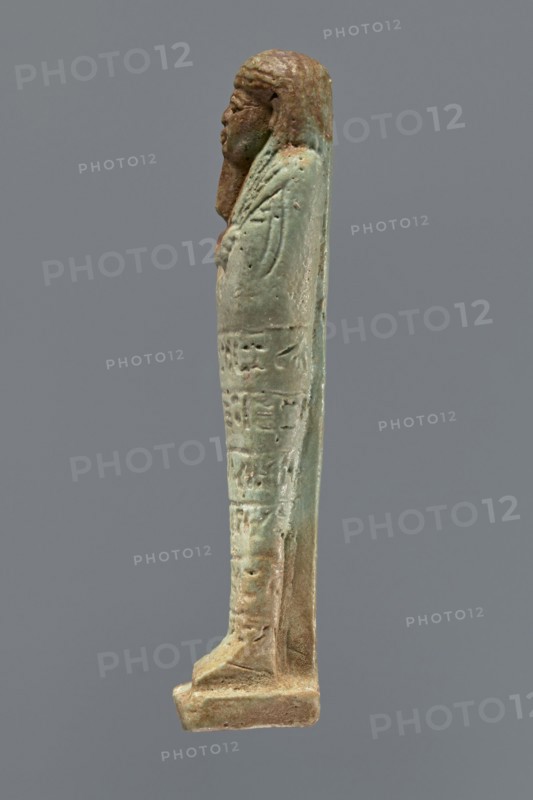Ushabti, mummy-shaped figurine, funerary gift, hoes. Mummy-shaped figurine standing on a base, supported by a pillar/pilaster at the back. Arms crossed on the chest, only the hands protrude from under the shroud, placed at the same level, close together, holding hoes in both hands. On the back, a basket is slung on a single string over the left shoulder. Engraved tools. On the head, a three-part wig styled in narrow strands. Long tapering beard, curled at the bottom and braided. Square face with almond-shaped eyes, pronounced eyebrows, wide nose, and prominent lips in a slight grimace. Engraved inscription arranged in six lines around the figurine; illegible. 664–332 BC; Late Period, Egypt. Shabti; height 15.5 cm., A slender standing figurine viewed in profile, with a sculpted head and stylized hair in brown tones and a body in greenish-blue and brown patina; the figure appears wrapped in a long garment with subtle raised details and surface texture, showing worn and rough areas, and i...