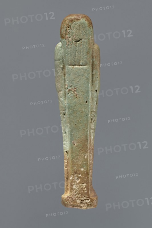 Ushabti, Mummy-shaped figurine, standing on a base, supported by a pillar/pilaster at the back. Arms crossed on the chest, only the hands protrude from under the shroud, placed at the same level, close together. Hoes in both hands. On the back, a basket is slung on a single string over the left shoulder. Engraved tools. On the head, a three-part wig styled in narrow strands. Long, tapering beard curled at the bottom and braided. Square face with almond-shaped eyes, pronounced eyebrows, a wide nose, and prominent lips in a slight grimace. Engraved inscription arranged in six lines around the figurine; illegible text. 664–332 BC; Late Period, Egypt. Shabti; height 15.5 cm., A narrow vertical statuette with a rounded top and flared base, shown against a plain gray background, displaying a weathered surface with a mix of pale green, turquoise, brown, and beige tones; the figure has simplified, elongated form with subtle raised ridges suggesting arms along the sides, pitted and rough areas,...