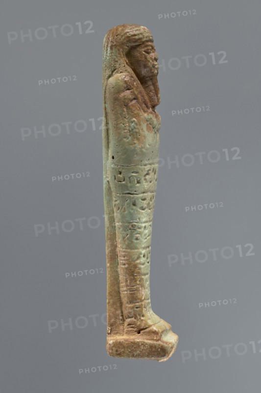 Ushabti, Mummy-shaped figurine, funerary shabti, 664–332 BC Late Period, Egypt. Standing on a base, supported by a rear pillar. Arms crossed on the chest with only the hands protruding from under the shroud, holding hoes in both hands. A basket is slung on the back over the left shoulder on a single strap. Engraved tools. Three-part wig styled in narrow strands. Long tapered beard, curled and braided at the bottom. Square face with almond-shaped eyes, pronounced eyebrows, wide nose, and prominent lips in a slight grimace. Engraved inscription arranged in six lines around the figurine; text illegible. Height 15.5 cm., A slender statuette of a standing human figure shown in profile with a long wig or headdress and a beard, rendered in a weathered greenish-blue and brown patina; the figure’s arms are held close to the body and the torso and lower body show incised horizontal bands and shallow carved details; the base is rectangular and the overall surface is pitted and worn with a mix of ...