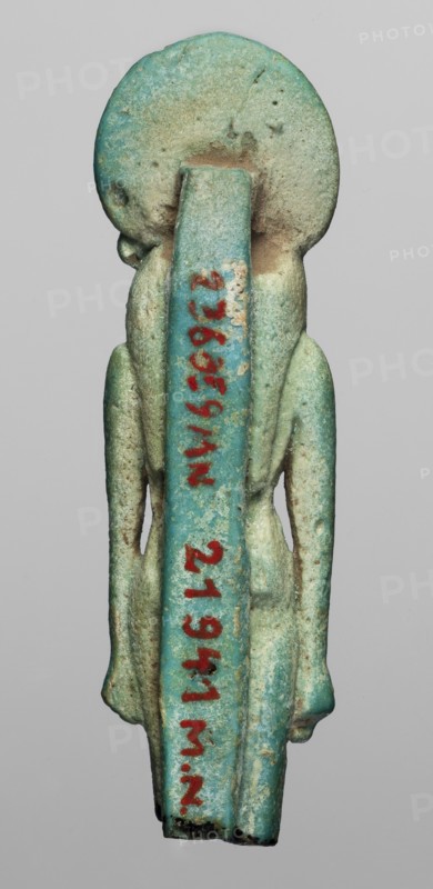 Amulet — figurine of a lion-headed goddess Sachmet?, Figurine of the goddess Sachmet with a lion's head topped with a solar disc with a uraeus, in a tripartite wig and a long narrow robe. The lower legs are missing, but the figure seems to be walking, with its arms lowered. On the back pillars. 664–332 BC; Late Period, Egypt. Amulet; height 5.8 cm, width 1.7 cm., A small vertical statuette photographed against a neutral gray background, showing the back of a standing human-like figure with an elongated straight back support and a large circular head; the surface has a weathered, grainy texture with areas of wear and small pits, and the predominant colors are turquoise and aqua with patches of pale green, sandy beige, and spots of darker green and brown; the figure has simplified arms hanging alongside the body and a slightly rounded base, and there is a vivid red-painted vertical marking along the back support that contrasts with the surrounding blue-green tones.