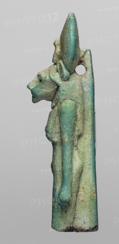 Figurine amulet of the goddess Sachmet lion-headed, possibly Bastet?, wearing a tripartite wig and a long narrow robe, topped with a solar disc with a uraeus. The lower legs are missing; the figure appears to be walking with arms lowered. Back pillars. Late Period, Egypt, 664–332 BC. Material: amulet; height 5.8 cm, width 1.7 cm., A slender vertical figurine in shades of turquoise, teal, pale green, and sandy beige shows a stylized anthropoid profile with an elongated head and pronounced snout, a rounded ear or headpiece, and a visible arm folded along the body; surface texture appears worn and pitted with areas of darker green and small patches of brownish discoloration, and there is a circular hole near the upper rear of the piece.