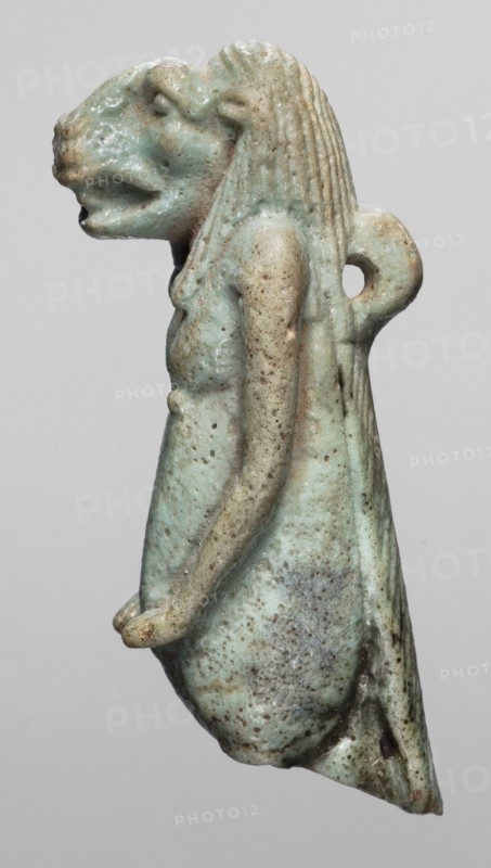Amulet - Toeris, repossession Radom, Toeris myth., Egyptian deities, hippopotamuses, Egyptian religion, 664–332 BC; Late Period, Egypt, amulet, height 4.6 cm, width 1.5 cm, A small sculpted figure with a lion-like head and human torso seen in profile, rendered in a pale green-blue color with areas of light beige and brown spotting, showing textured hair with vertical ridges, a looped projection at the back, a partial arm extending forward with a rounded hand, a tapered lower body with rough pitted surface and darker speckling, and an overall worn, slightly glossy surface against a plain gray background.