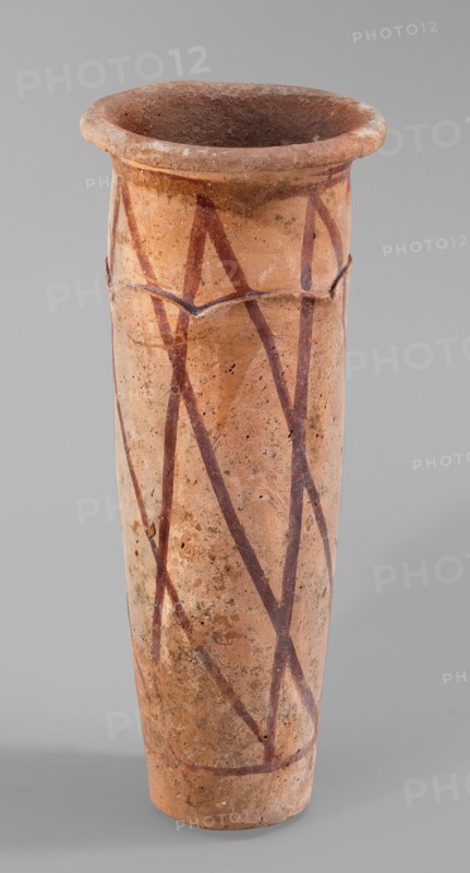 Cylindrical vessel with crosshatched decoration, painted ceramic with engraved decorations and geometric motifs. The vessel has a shape resembling a cylinder, slightly tapering towards the bottom, with a slightly convex bottom and a widely rolled-out rim. A plastic, wavy ornament runs 3/4 of the way up; it is arranged so the upper part of the vessel appears recessed relative to the lower. Surface covered with a light engobe and decorated with a painted diagonal grid. Attributed to unknown author, possibly B. Ruszczyc. c. 3200–c. 3050 BC; Predynastic Period, Naqada III Naqada phase IIIa2, from Abusir el-Melek. Vessel. Height 27.5 cm; width 11 cm; weight 1.028 kg., A tall cylindrical ceramic vessel with a flared rim, showing a worn, textured surface in shades of light brown and tan with darker brown and reddish-brown painted zigzag lines and spots; the interior appears darker brown, and the background is a neutral gray.