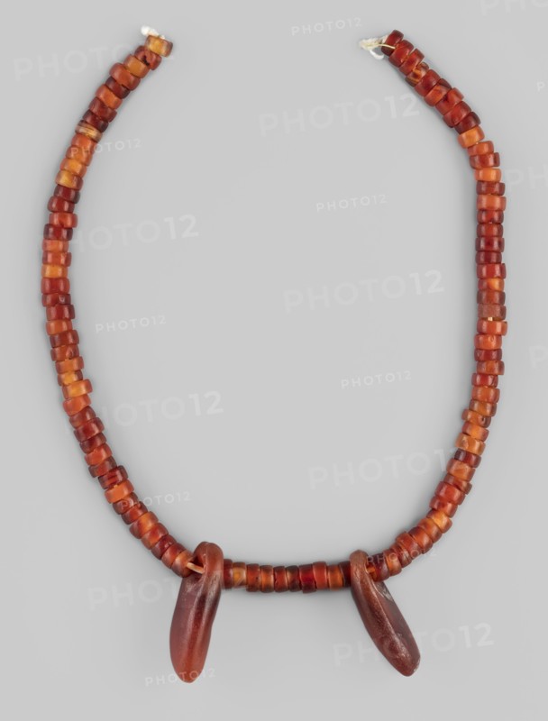 Necklace - fragment, cylindrical beads, Necklace of cylindrical beads interrupted by two pendants of elongated, irregular shape, in various shades of red, 5 thousand BC, Kadero grave no. 3, jewelry, height 25.2 cm, A beaded necklace arranged in a loose U shape against a light gray background, composed of a string of small cylindrical beads in shades of red, orange, and amber, with a few beads showing darker reddish-brown tones; two larger elongated pendant beads in darker reddish-brown hang from the lower center of the strand.