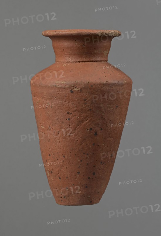 Vessel, Egyptian pottery, clay vessel, conical-shaped vessel, widest at approximately 3/4 of its height. Quite wide, almost horizontal shoulders—slightly sloping. Wide, short, funnel-shaped neck widening towards the spout. Wide spout, flat in the upper part, slightly rounded at the bottom, slightly asymmetrical. Flat, asymmetrical bottom. Surface leveled. Lower part finished to approximately 2/3 of its height with a knife or similar tool. Outer surface and inner surface of rim and neck covered with a layer of dark red engobe. Vessel color: dark red. Vessel complete; surface eroded in many places, numerous clay chips on the rim. Ca. 2686–ca. 2181 BC; Old Kingdom, Edfu. Vessel height 16 cm; diameter 8.6 cm rim, 10.7 cm body, 4.5 cm bottom., A single clay vessel with a rounded shoulder and a flared rim, shown centered against a plain gray background, has a rough textured surface with visible pitting and small dark speckles; the vessel is mainly terracotta red with patches of darker brown ...