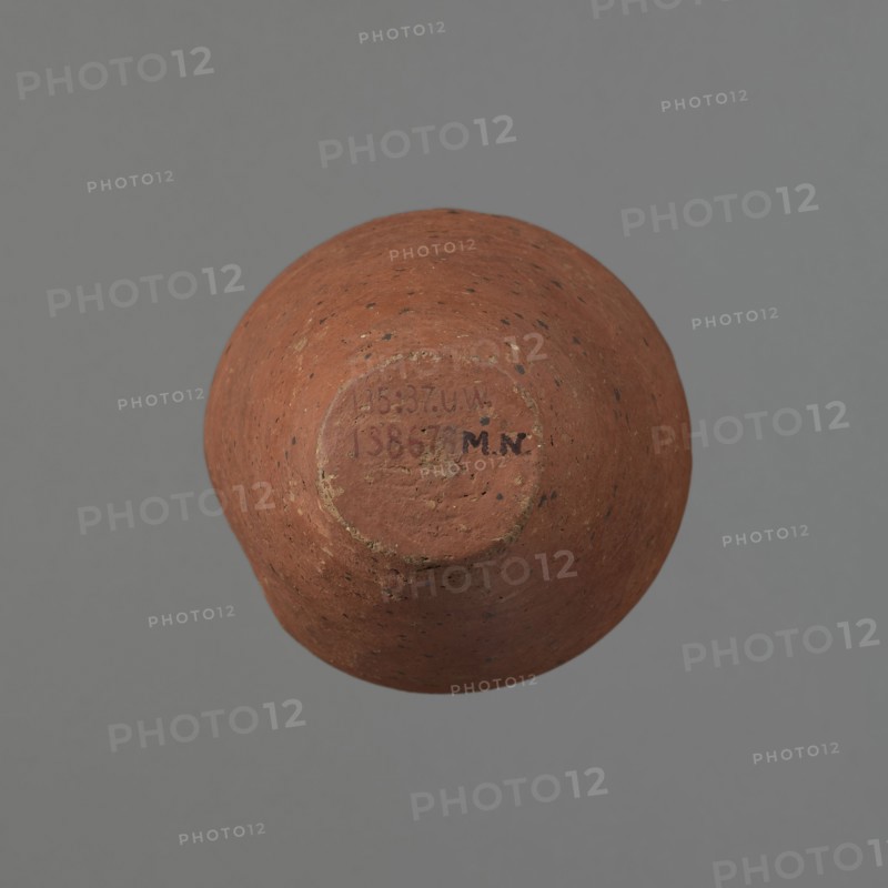 Vessel, Egyptian pottery, ceramics, clay vessel. Conical-shaped vessel, widest at approximately 3/4 of its height. Quite wide, almost horizontal shoulders slightly sloping. Wide, short, funnel-shaped neck, widening towards the spout. Wide spout, flat in the upper part, slightly rounded at the bottom, slightly asymmetrical. Flat, asymmetrical bottom. Surface leveled. Lower part finished to approximately 2/3 of its height with a knife or similar tool. Outer surface and inner surface of the rim and neck covered with a layer of dark red engobe. Vessel color dark red. Vessel complete; surface eroded in many places, numerous clay chips on the rim. Old Kingdom, ca. 2686–2181 BC; provenance Edfu. Vessel height 16 cm; diameter 8.6 cm rim, 10.7 cm body, 4.5 cm bottom., A rounded terracotta object with a rough, speckled surface in shades of reddish-brown and warm orange sits against a flat gray background, showing a recessed circular base area with worn edges and traces of a darker brown patina; ...