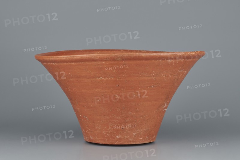 Deep, cup-shaped bowl with a slightly convex bottom and sides arching upwards; straight, flat, flaring rim. Made of Nile clay NB1 ? according to the Vienna System, covered with an engobe containing red ochre and thoroughly polished. Color: red. Well-preserved with slight clay damage on the rim. Comparative attributions: cited parallels to pottery and typologies from the 4th–6th Dynasties / Old Kingdom. Vessel dimensions: height 15.1 cm; lip diameter 27.6 cm; bottom diameter 11.8 cm; weight 1.3 kg., A single terracotta-colored clay vessel with a flared rim and conical body sits against a smooth gray background; the surface of the vessel shows variations in the terracotta hue with lighter and darker patches and some small worn or rough spots, and the scene includes shades of gray in the backdrop and base area.