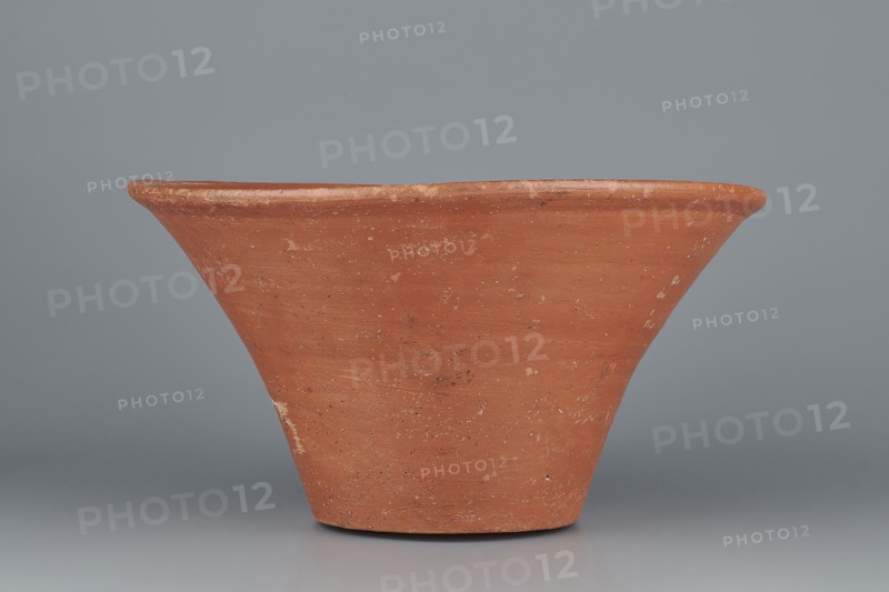 Deep bowl. Cup-shaped clay bowl with a slightly convex bottom and sides arching upwards, straight flaring rim flat on top. Made of Nile clay NB1 ? with an engobe containing red ochre, thoroughly polished. Color: red. Well-preserved with slight clay damage on the rim. Function: vessel. Dimensions: height 15.1 cm; lip diameter 27.6 cm; bottom diameter 11.8 cm; weight 1.3 kg. Attributed date/style: Old Kingdom, 4th–6th Dynasty c. 2494–c. 2181 BC. Comparative parallels: parallels noted with Mycerinus and Giza Necropolis pottery and late 6th-dynasty and Early Bronze Age III types. Maker: unknown., A wide, flared terracotta vessel with a rough, matte surface in shades of orange-brown and reddish-brown, showing lighter beige and white spots and scuffs, set against a smooth, cool gray background; the vessel tapers toward a narrower base and has a slightly rounded rim with visible texture variations.