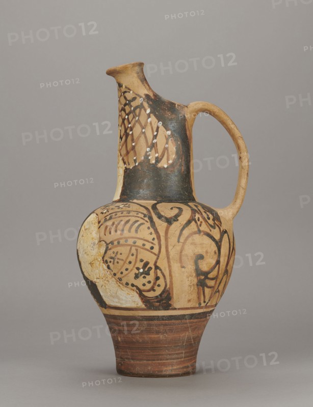 Red-figure oinochoe by the Pennsylvania Torcop Painter, Classified Gallo-Classic Scenography; red-figure ceramic wine vessel depicting mythological scenes and women; 2nd half of the 4th–3rd century BC c. 300–280 BC, to be verified; Etruria place of origin; vessel; height 31.5 cm, width 16.2 cm, depth 15.6 cm, weight 0.94 kg, A ceramic jug with an elongated neck, curved handle, and rounded body standing on a conical foot; the surface shows painted decorative patterns and areas of worn or flaked paint. The visible colors include beige and tan on the clay body, black and dark brown used for painted motifs and a darker area on the neck, white applied in small spots and patches, and reddish-brown bands around the foot. The decoration features flowing curved lines, spiral-like shapes, dotted accents, and a lattice-like pattern on the upper neck, with some areas where the paint is chipped or missing revealing the underlying lighter clay.
